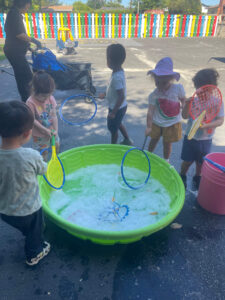Five preschool children playing with a tub of water outdoors, enjoying sensory play and outdoor learning activities.