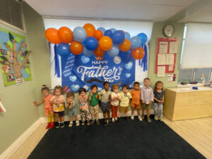 Young children posing in front of a Happy Father's Day flag against the wall of a classroom.