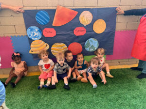 Toddlers posing proudly in front of a solar system poster they created during a preschool daycare science activity.