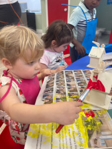 Toddlers painting a wooden house at a blue table during an arts and crafts activity at daycare.