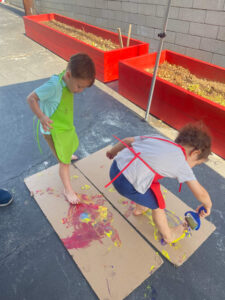 Two preschool boys painting a cardboard outside on the floor using their feet during an art activity.