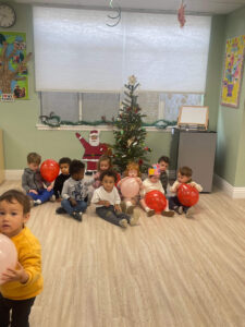 Young children sitting on the floor in a festive daycare classroom with a Christmas tree during the holiday season.