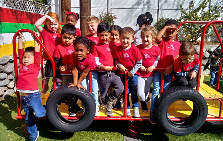 Preschool children posing for a photo on the playground, all wearing matching red Firehouse School shirts.