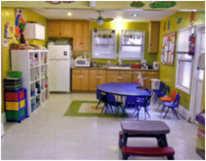 Firehouse School preschool classroom with green walls, a blue activity table, cubbies for storage, and a child-friendly fridge.