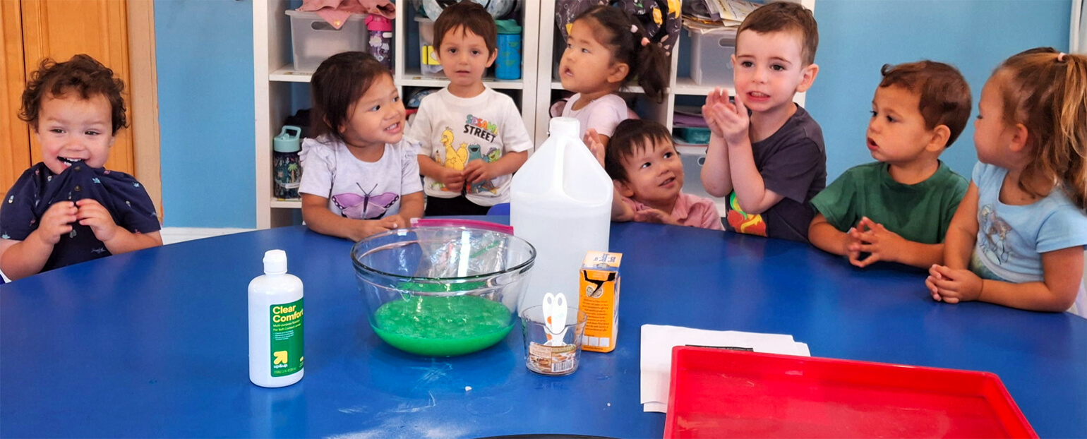 Preschool children smiling and gathering around a blue table, preparing for a fun early childhood learning activity.