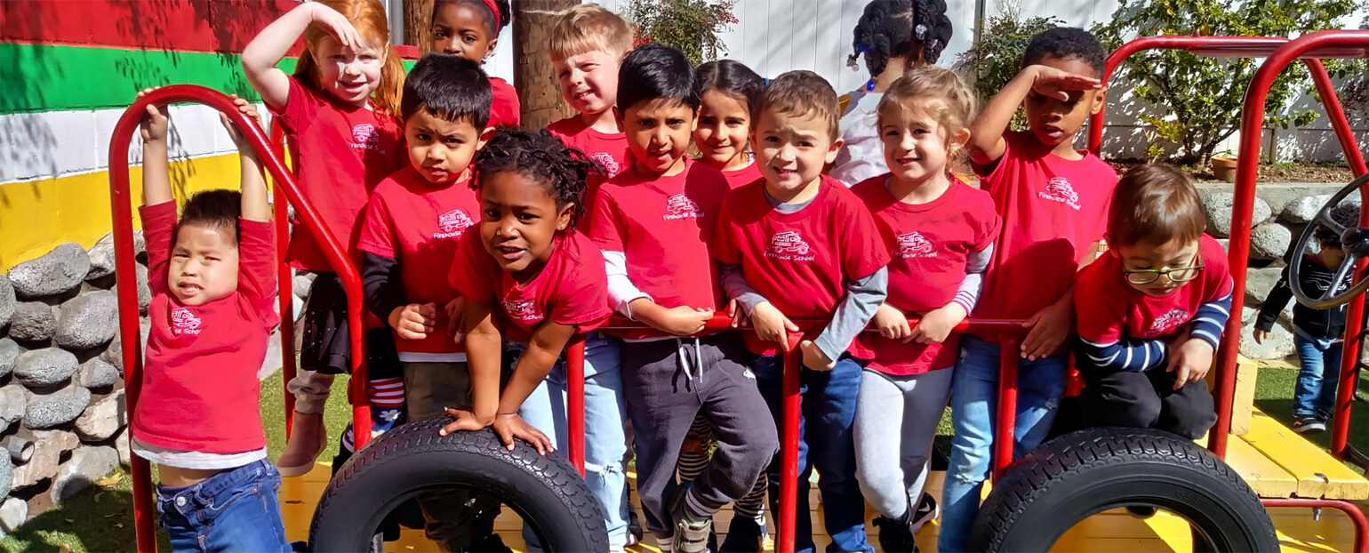 Preschool children posing for a photo on the playground, all wearing matching red Firehouse School shirts.