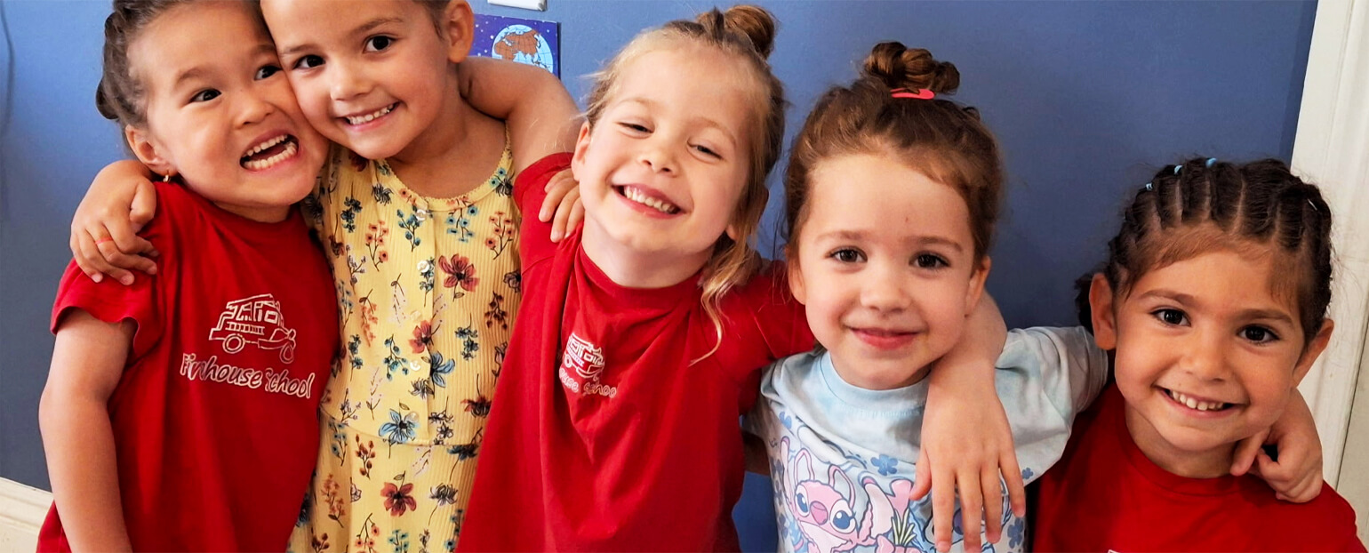 Five children at daycare smiling and posing for a photo against a blue classroom background.